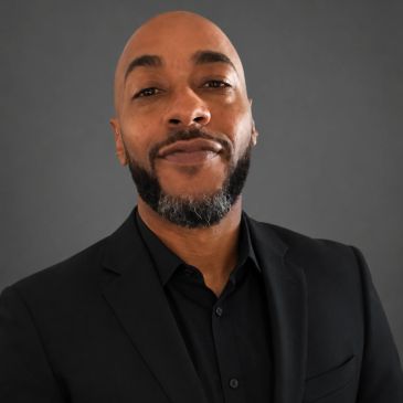 Confident man in a black suit with a trimmed beard and bald head.