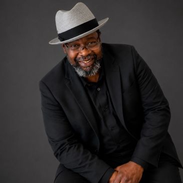 Smiling man in a gray fedora and black suit against a dark backdrop.