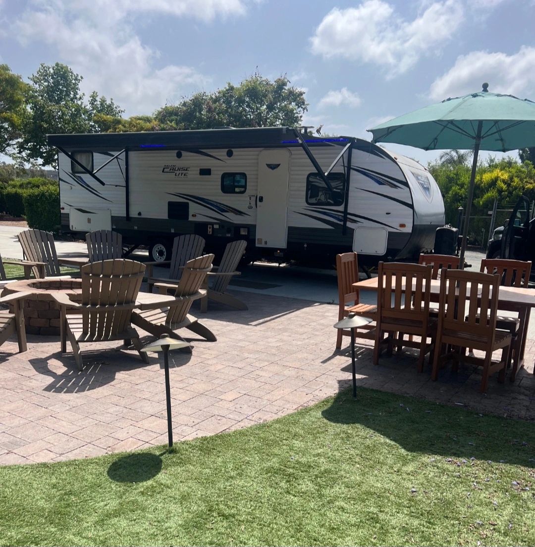Outdoor patio with chairs, dining table, and a parked travel trailer under a partly cloudy sky.