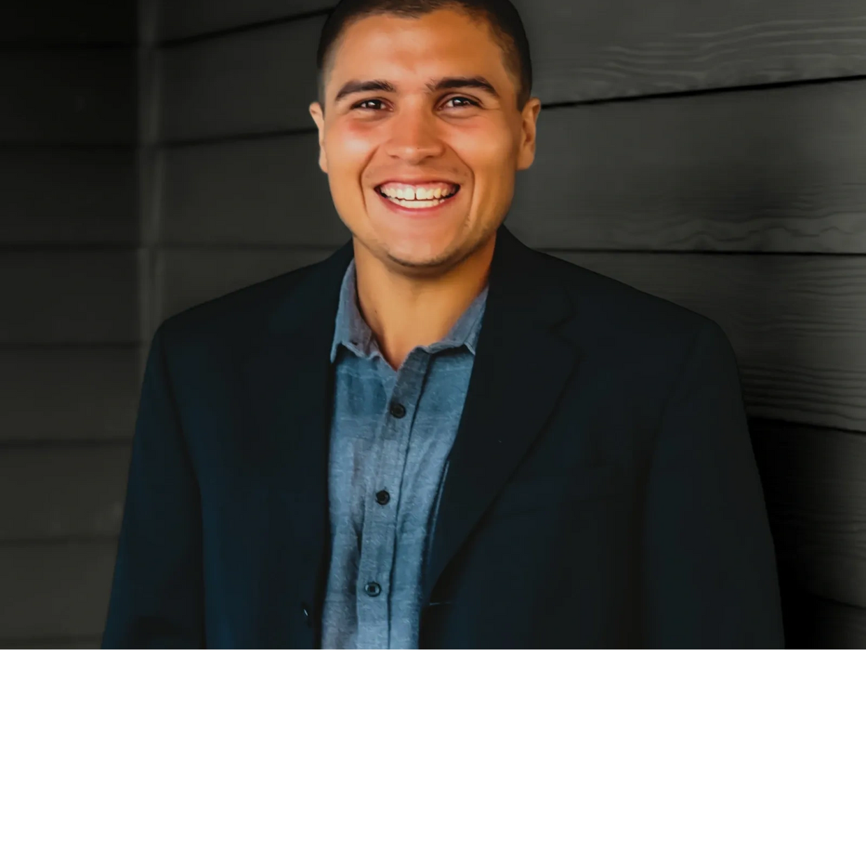 A young man in a blazer smiling brightly against a dark wooden background.