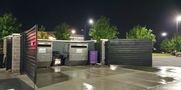 Nighttime scene of dumpsters enclosed by walls in a parking lot area.
