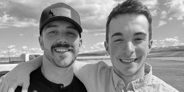 Two smiling men posing outdoors with blue sky and clouds behind them.