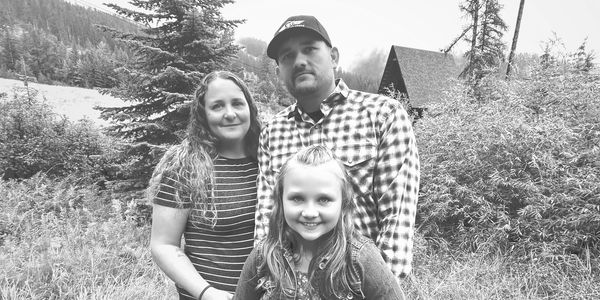 A family of three posing outdoors in a lush green field with trees and a cabin in the background.