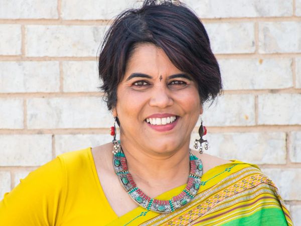 Smiling woman in a yellow and green sari with traditional jewelry.