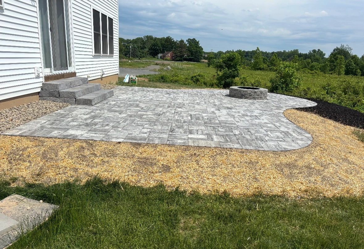 Newly constructed stone patio with firepit and steps beside a white house.