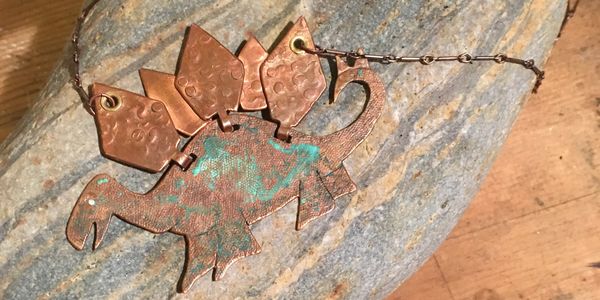 A copper hand-wrought playful Stegosaurus necklace with a green patina and leather band.