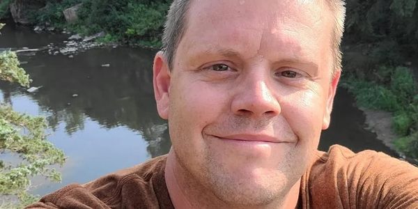 A man taking a selfie near a river with trees in the background.