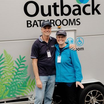 Two people smiling in front of an Outback Bathrooms trailer.