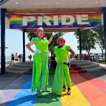 Two people in bright green outfits pose under a rainbow PRIDE banner outdoors.