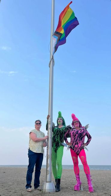 Two drag performers and a woman raising a pride flag on a beach under clear sky.