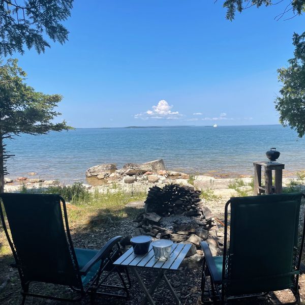 Two camp chairs are in front of a fire pit. there is a table between them. A lake is nearby