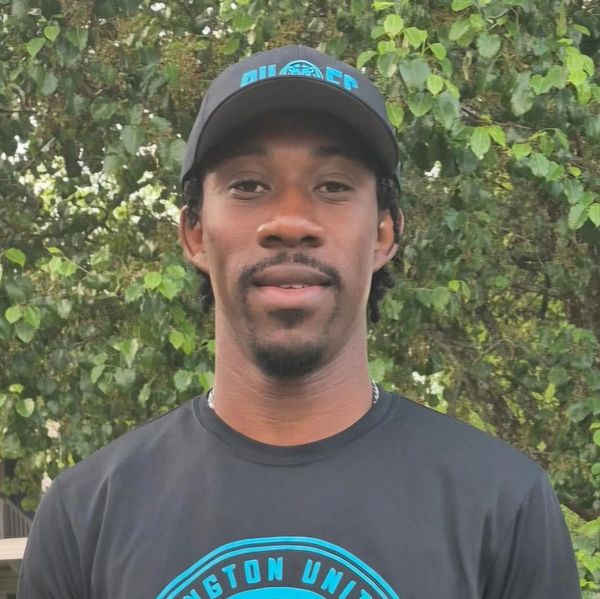 Man in a black cap and t-shirt standing outdoors with greenery in the background.