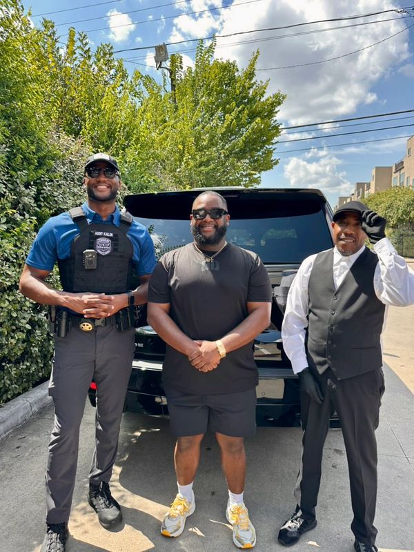 Three men standing and smiling in front of a black vehicle on a sunny day.