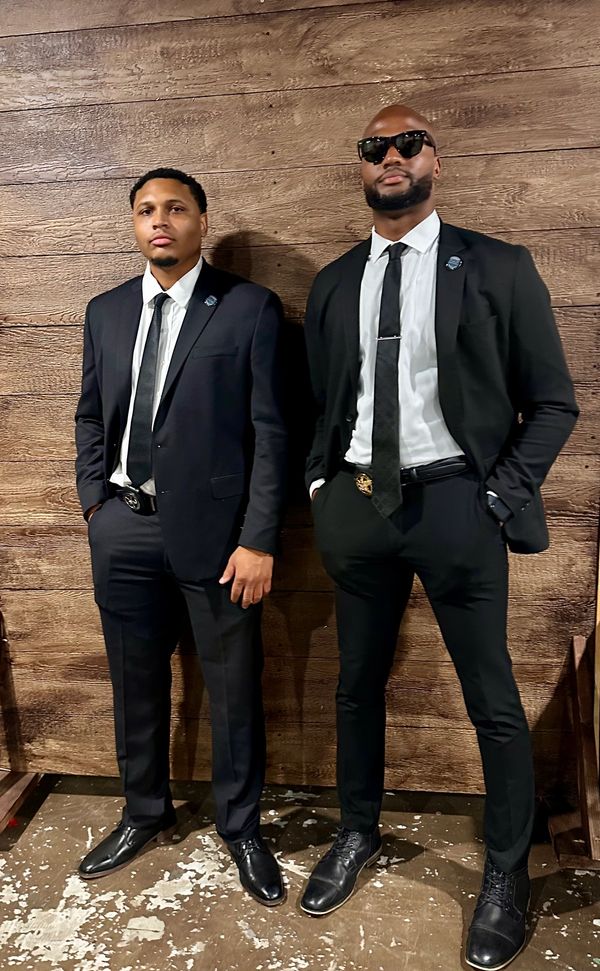 Two men in black suits and ties standing against a wooden wall.