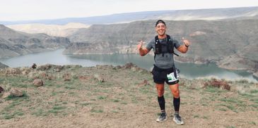 Smiling runner gives thumbs up on a scenic trail with river and cliffs in the background.