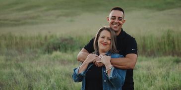 Couple embracing happily in a grassy field with hills in the background.