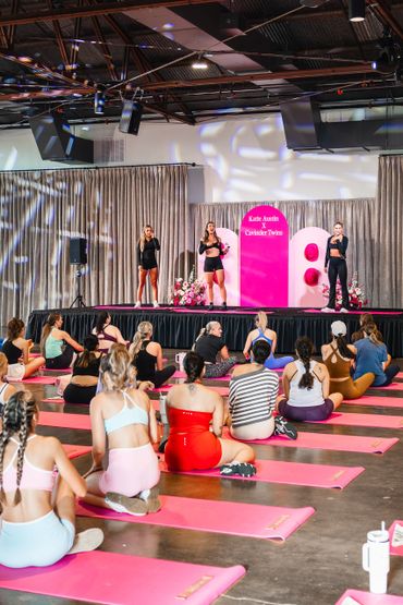 Group fitness class with participants on pink mats following instructors on stage.