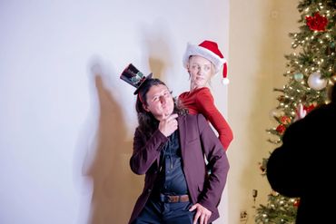 Two people posing humorously with festive hats near a Christmas tree.