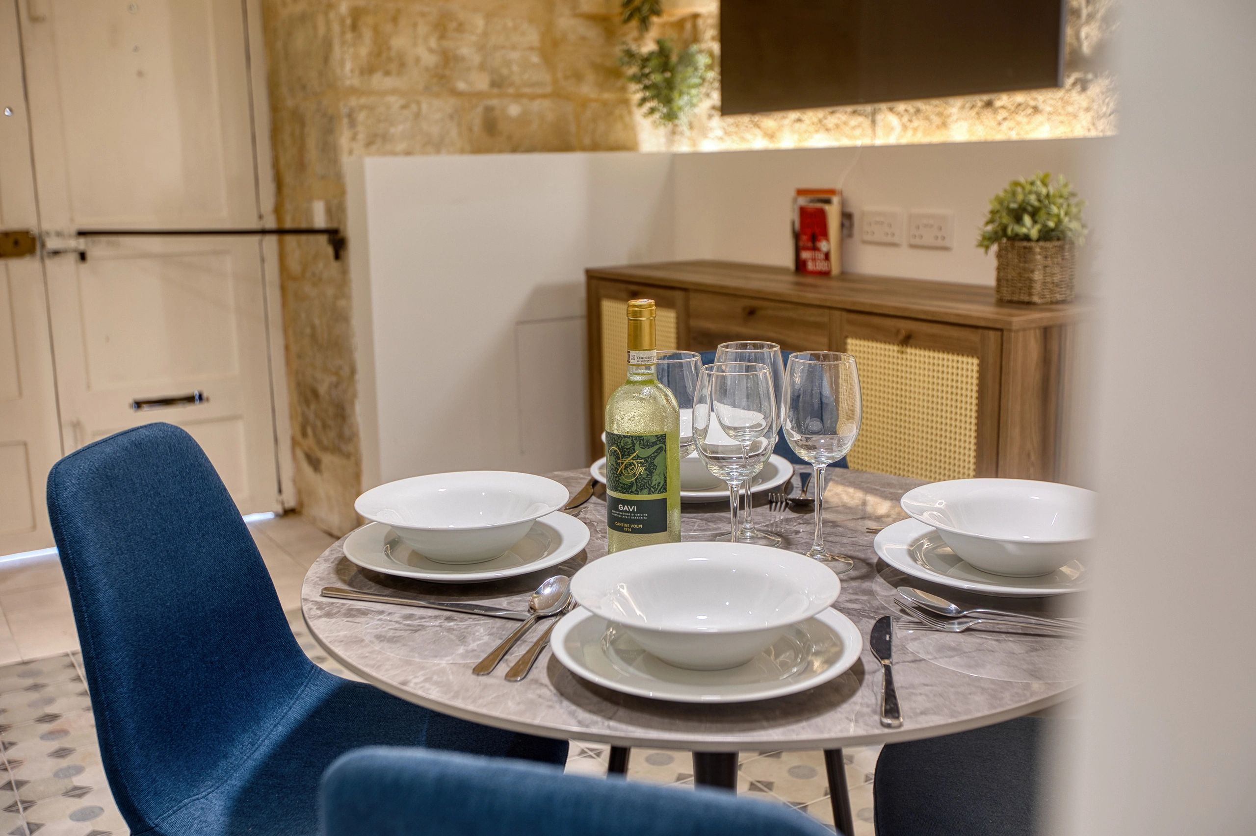 Elegant dining table set with white plates, wine glasses, and a bottle, surrounded by blue chairs.