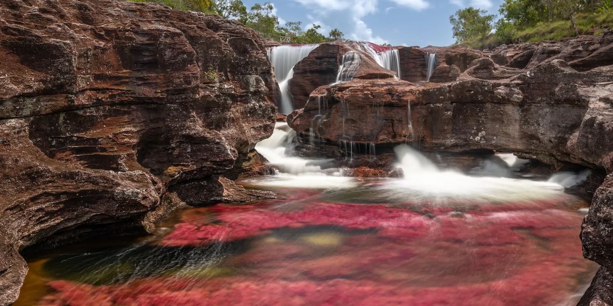 Waterfall flowing over rocky terrain with vibrant red aquatic plants in the water.
