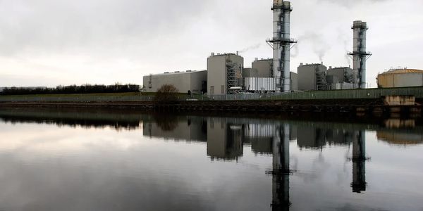 Industrial plant with smokestacks reflecting on a calm water body under a cloudy sky.