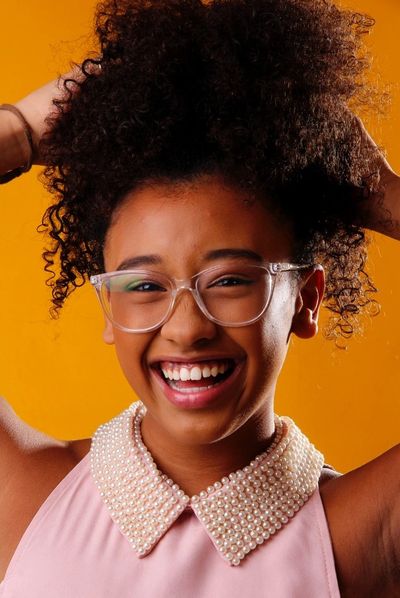 Smiling woman with curly hair, glasses, and a pearl-collared top against an orange background.