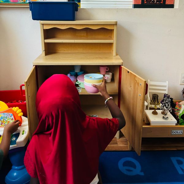 Child in red hijab playing with toy kitchen set in a classroom.