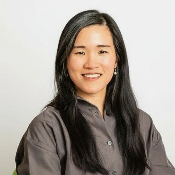 Smiling woman with long black hair seated on a green chair against a plain background.