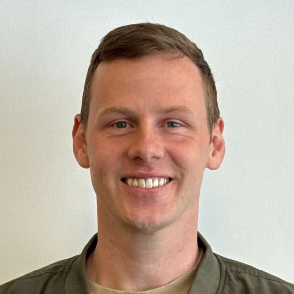 Smiling young man wearing a green jacket and beige shirt against a plain white background.