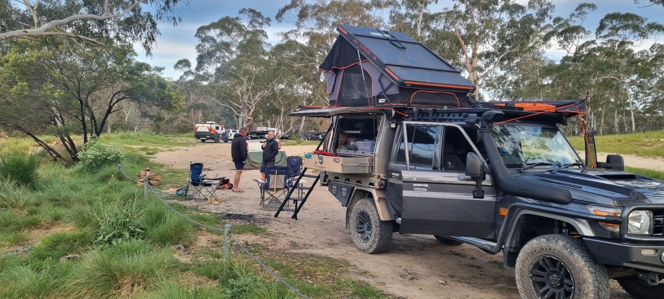 Two men camping near a black off-road vehicle with rooftop tent in a forested area.