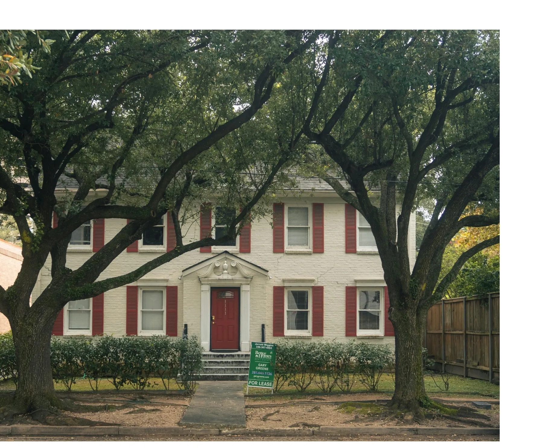 Two large trees frame a white brick house with a red door and shutters.