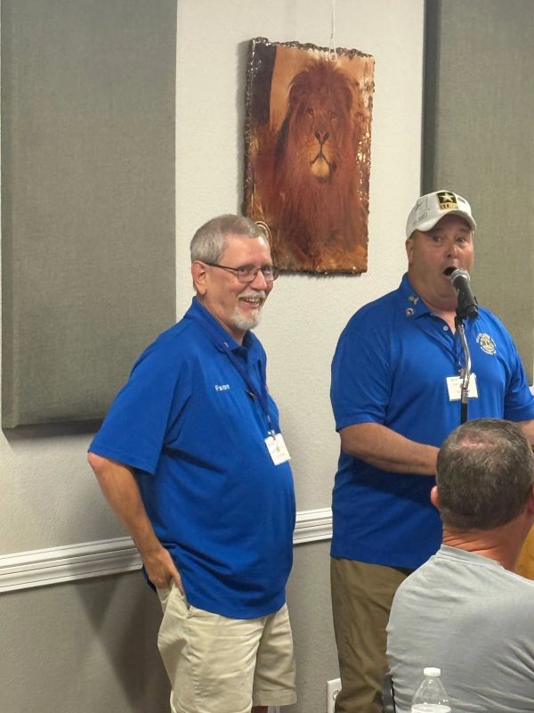 Two men in blue shirts stand in a room with a lion painting.