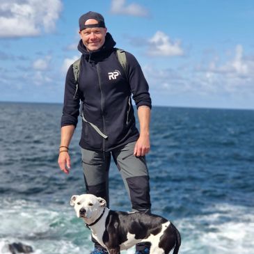 Man with dog on rocky shore under blue sky.