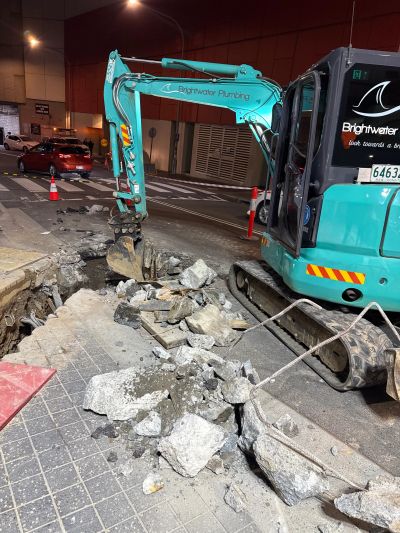 Brightwater Plumbing excavator breaking concrete on a street at night.