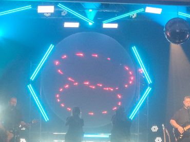 Band performing on stage with vibrant neon lights and circular LED display.