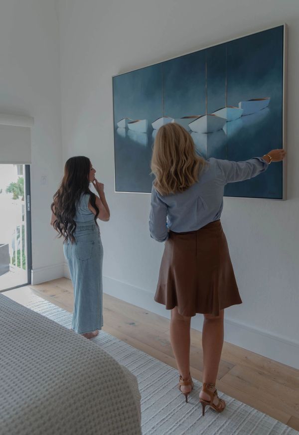 Two women admire a large painting of boats on a calm blue sea in a modern room.