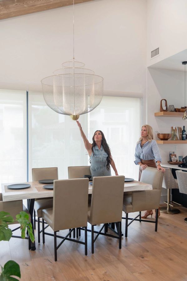 Two women in a modern dining room, one reaching towards a large hanging light fixture.