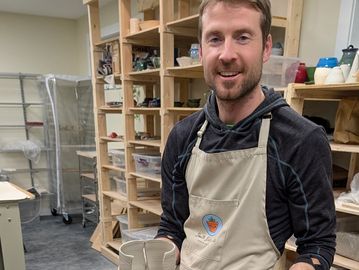 Man in apron holding ceramic cups in a pottery studio.