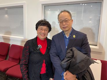 Two elderly individuals smiling indoors with red chairs in the background.
