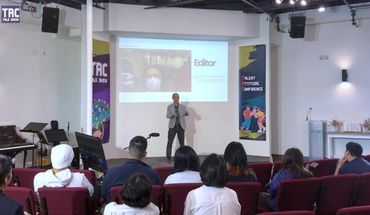 A speaker presents on stage to an attentive audience in a modern conference room.