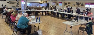 A community meeting with people seated around tables in a large room.