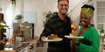 Two people holding plates of food and smiling in a kitchen.