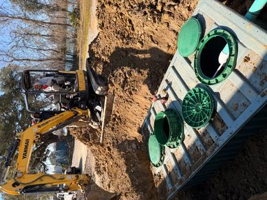 Construction workers operating a small excavator near green plastic pipes in a dirt trench.