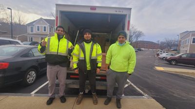 Three men in yellow safety jackets stand in front of a moving truck in a parking lot.