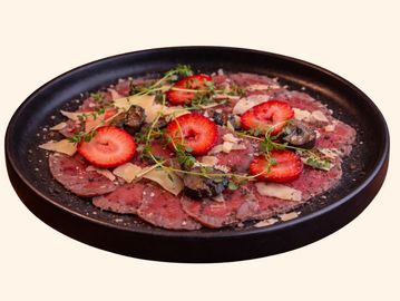 Gourmet beef carpaccio with strawberries, mushrooms, herbs, and cheese on a black plate.