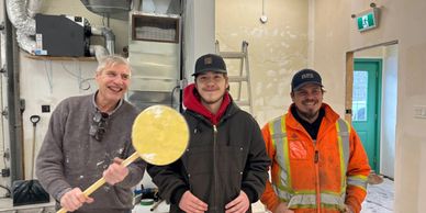 Three men smiling, one holding a pizza peel in a workshop.