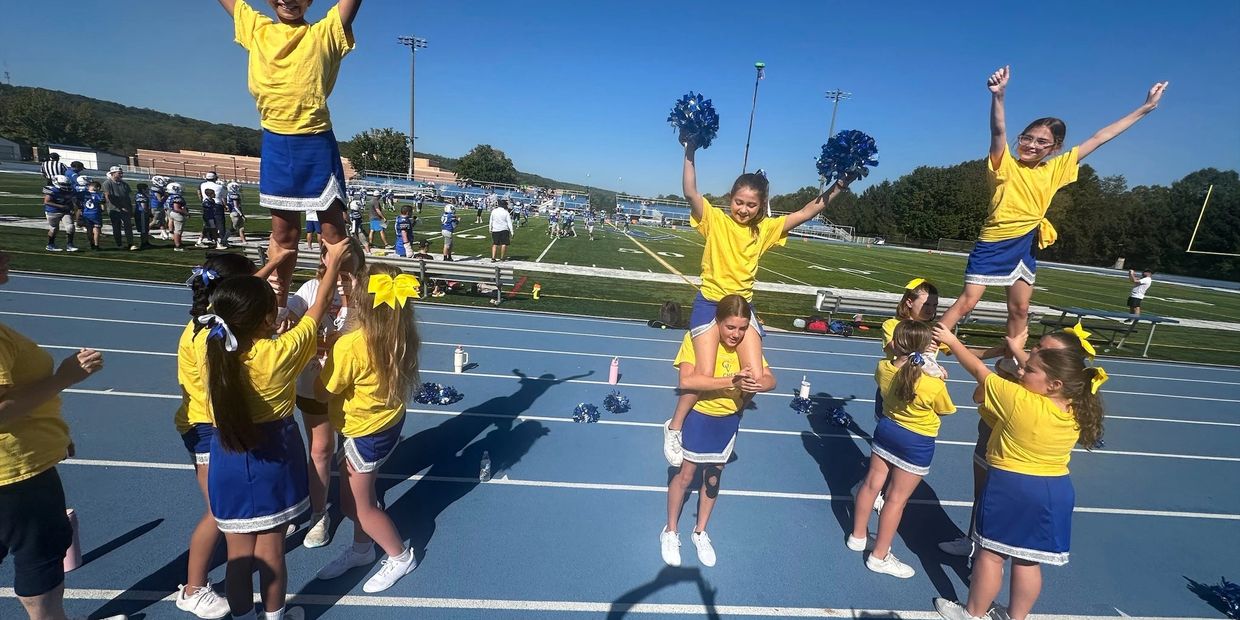 Cheerleaders in yellow tops and blue skirts perform stunts on a sunny football field.