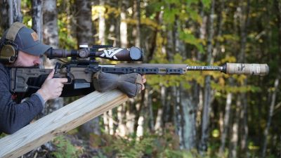 Man aiming a rifle with scope outdoors off a PRS Tank Trap Barricade.