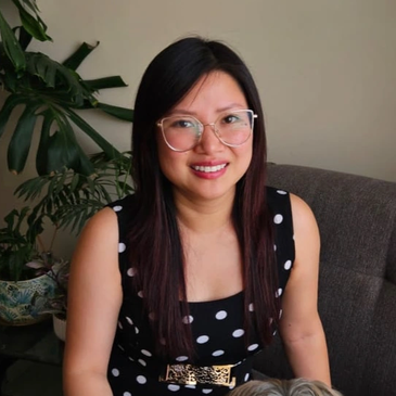 Smiling woman with glasses in a black and white polka dot dress sitting on a couch.