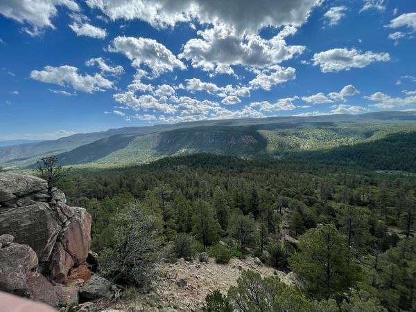 Expansive forest and mountain landscape under a partly cloudy blue sky.